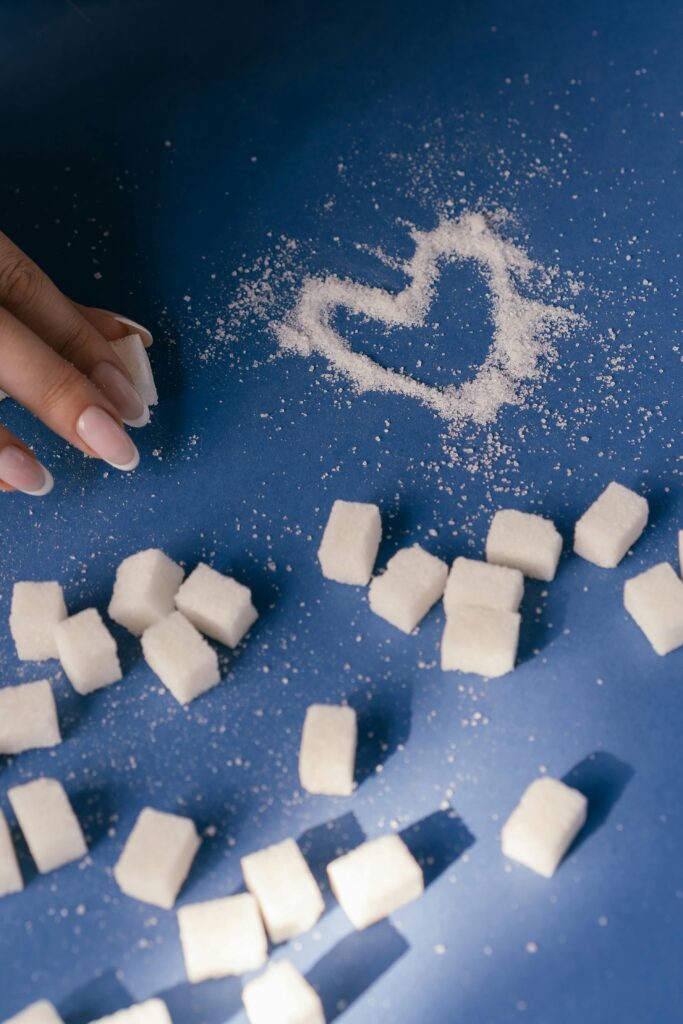 Top view of sugar cubes and a heart drawn in sugar on a blue surface with a hand nearby.